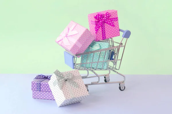 Pequeñas cajas de regalo de diferentes colores con cintas en el carrito de la compra sobre un fondo pastel violeta y azul —  Fotos de Stock