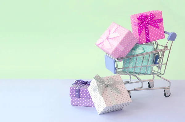 Pequeñas cajas de regalo de diferentes colores con cintas en el carrito de la compra sobre un fondo pastel violeta y azul —  Fotos de Stock