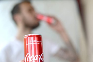Caucasian man drinks Coca-Cola drink in garage interior and male hand presents one Coca cola red can in focus clipart