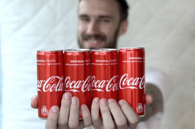 Smiling man holding many non-alcoholic Coca-Cola aluminium tin cans in garage interior clipart
