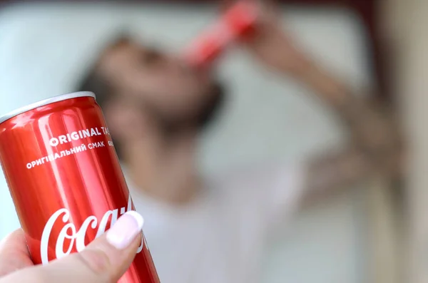 Kaukasischer Mann trinkt Coca-Cola-Drink im Garageninneren und männliche Hand präsentiert eine rote Dose Coca-Cola im Fokus — Stockfoto