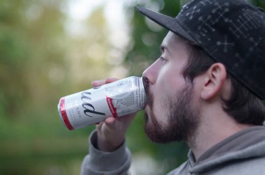 Genç sakallı adam Budweiser bira içiyor alüminyum kutuda yeşil ağaçlarda bulanık arka planda