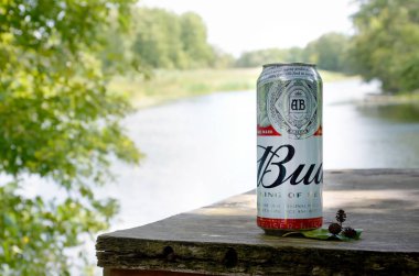 Budweiser Bud bira kutusu nehir kenarındaki eski ahşap masa ve yeşil ağaçların arkasında.