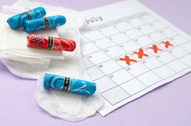 Adet takvimindeki regl pedleri ve tamponlar leylak arka planında kırmızı haç işaretli.