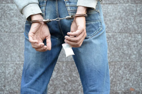Арестованный наркоторговец в полицейских наручниках с маленьким пакетом героина на фоне темной стены — стоковое фото