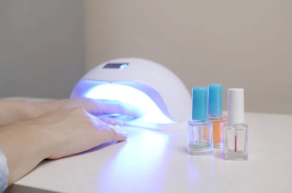 Three colorless transparent nail polish bottles in background of female hands in gel uv led nail white lamp for drying manicure