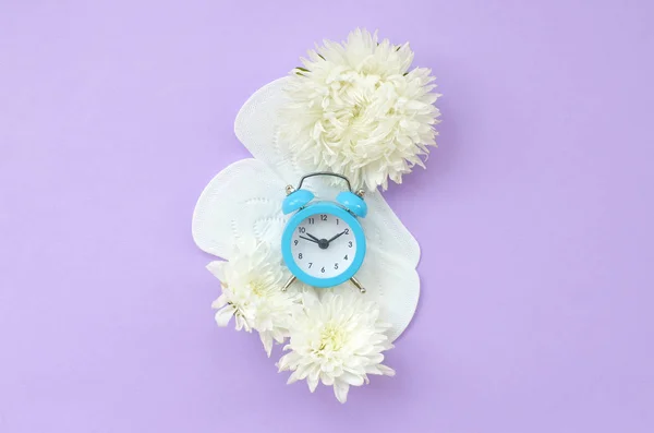 Blauer Wecker und weiße Blumen liegen auf Menstruationspolstern auf pastellfliederfarbenem Hintergrund — Stockfoto