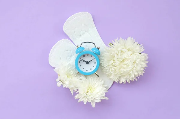 Relógio de alarme azul e flores brancas encontra-se em almofadas menstruais em fundo lilás pastel — Fotografia de Stock