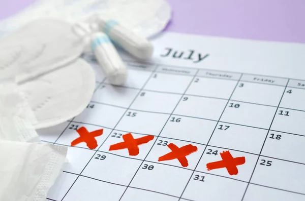 Almohadillas y tampones menstruales en el calendario del período de menstruación con marcas de cruz roja se encuentra en el fondo lila — Foto de Stock