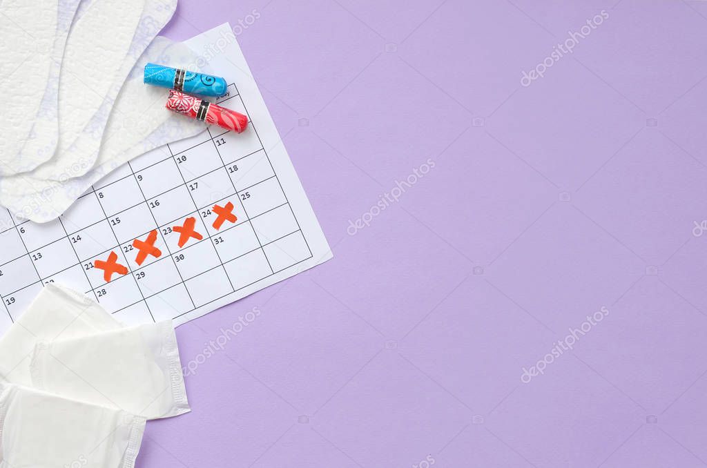 Menstrual pads and tampons on menstruation period calendar flat lay on lilac background