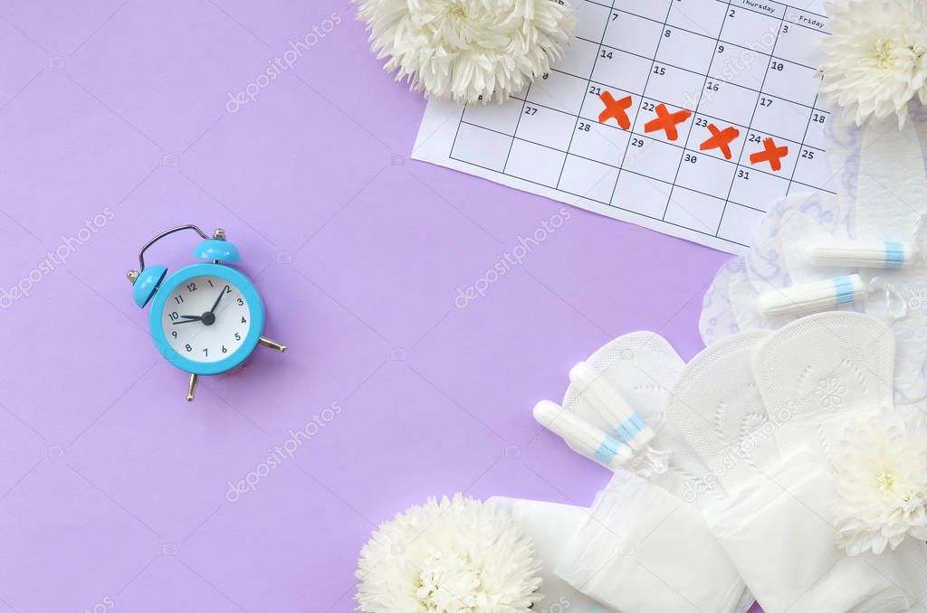 Menstrual pads and tampons on menstruation period calendar with blue alarm clock and white flowers