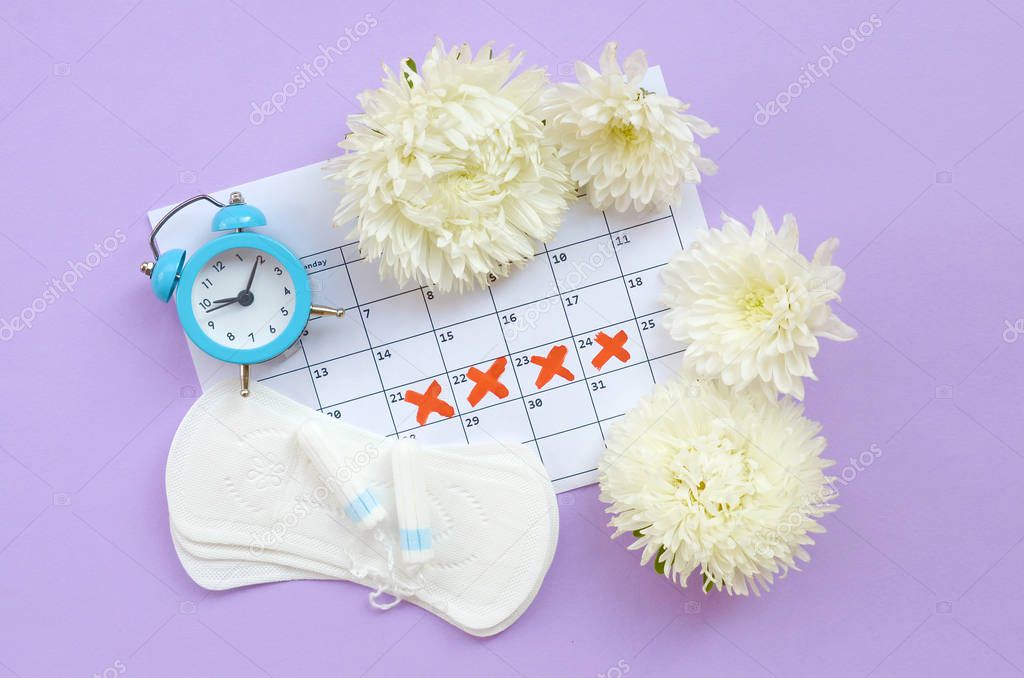 Menstrual pads and tampons on menstruation period calendar with blue alarm clock and white flowers