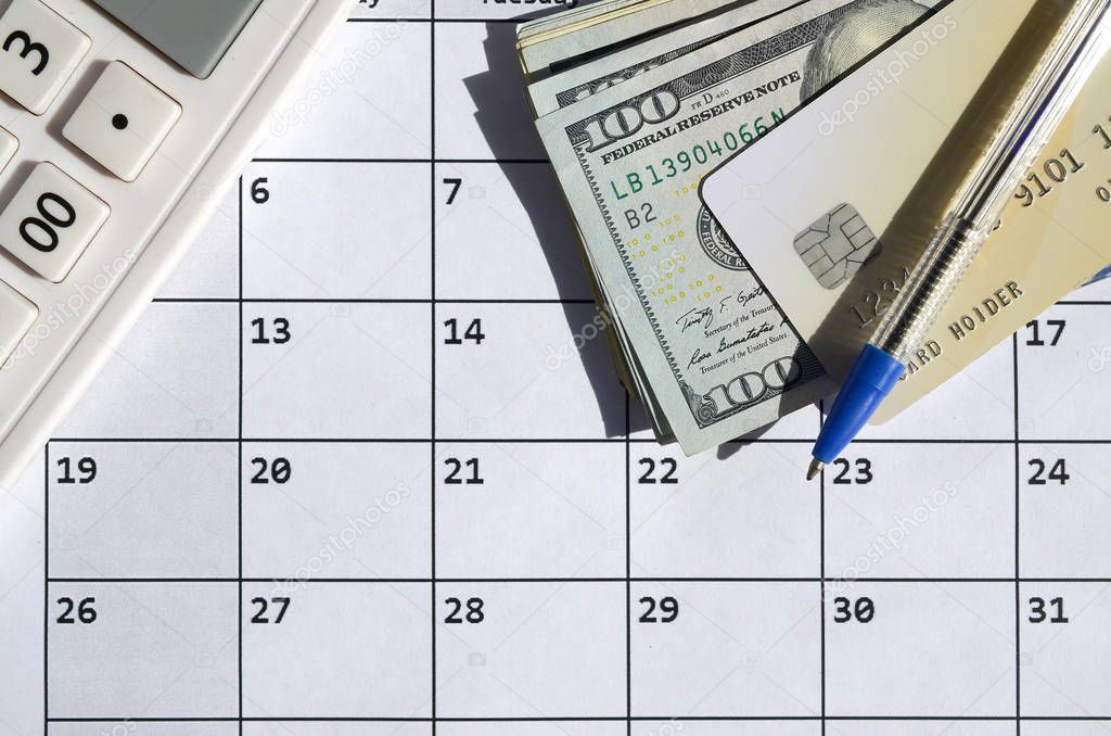 Pen and credit card on many hundred US dollar bills and calculator on calendar page close up
