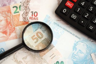 Brezilya reais banknotları ve büyüteçli hesap makinesi. Mevcut Brezilya gerçek para faturaları ve loupe ile calc büyük miktarda masada yatıyor. Brezilya 'da vergilendirme ve para konsepti
