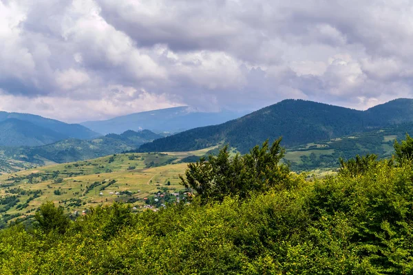 Un cielo gris nublado que se extiende sobre un pequeño pueblo rural a la sombra de las enormes montañas verdes de los Cárpatos — Foto de Stock