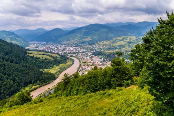 Una pintoresca vista de un pueblo rural y un río rápido sobre el fondo de altas montañas verdes —  Fotos de Stock