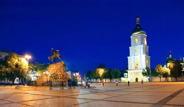 Evening lights of the city in a large square with standing in the middle of it a pamper with a rider on horseback. Opposite him is a church with light in the windows and a dome with a cross. — Stock Photo, Image