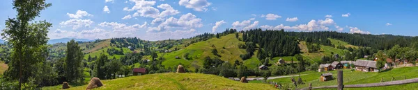 A charming panorama of mountainous green slopes on a summer day with rural houses scattered around the country with dry harvested hay on farms and white rare clouds floating on the horizon line. — Stock Photo, Image