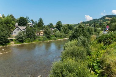 mavi bir bulutlu gökyüzü altında mesafe çok uzak bir yol ile yeşil ağaçlar ve çalılar özel kırsal konutlarında ve orman kaplı dağ arasında gizli boyunca akan bir nehir doruklarına. dinlenme yer