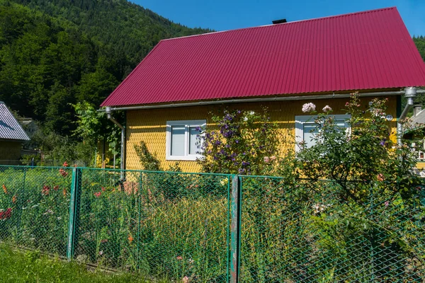 Casa amarela com um telhado vermelho e um belo canteiro de flores ao lado de uma cerca verde — Fotografia de Stock