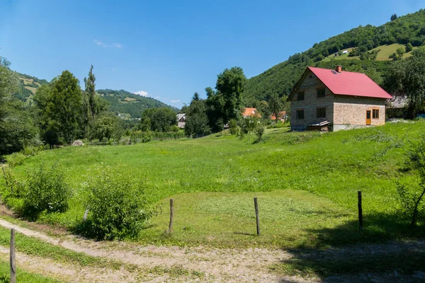 Uma estrada rural correndo ao lado do quintal com um gramado verde e uma casa em pé nas profundezas da colina . — Fotografia de Stock