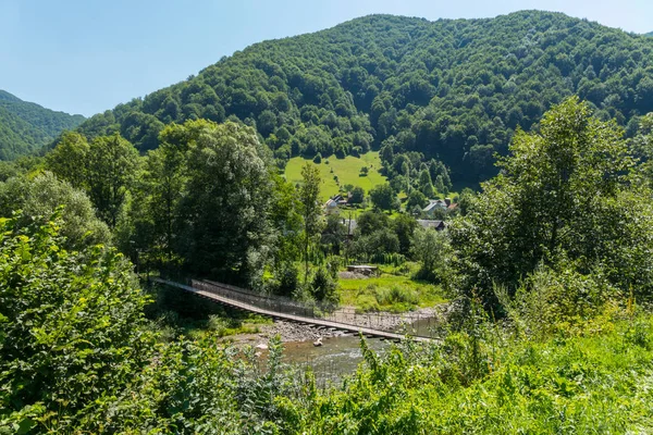 Nehrin yanında yeşil bir karşı kıyısında ayakta kalın ağaçları ile büyümüş bir eğim ile kırsal konutlarında yol trafiğe kapalı bir asma köprü.