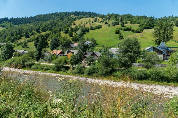 Die Dorfstraße mit gepflegten Häusern liegt am Fuße des Hügels mit grünen Hängen und der Strömung unterhalb des Flusses. — Stockfoto