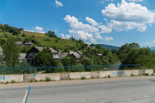 Aldeia em uma encosta de montanha verde atrás de uma estrada de asfalto em uma ponte através de um rio tempestuoso abaixo de um céu nublado azul. lugar de descanso e turismo — Fotografia de Stock