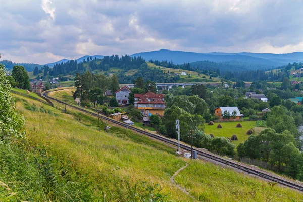 Die Eisenbahn fährt zwischen den Berghängen entlang des verhangenen Grüns des Dorfes vor dem Hintergrund nebelverhangener Berggipfel unter wolkenverhangenem Himmel. Ort der Ruhe und des Tourismus — Stockfoto