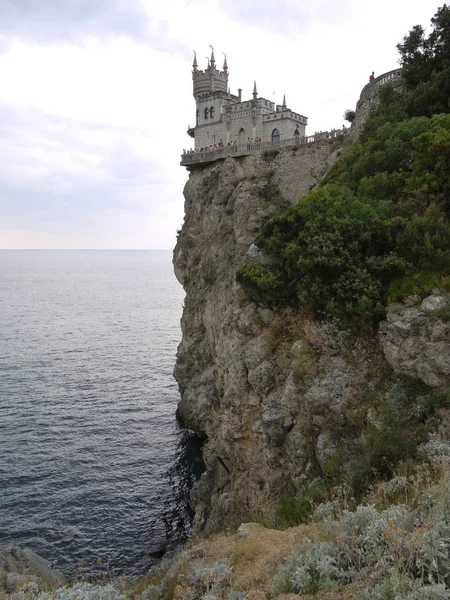 A castle with turrets located at the very edge of a cliff standing in the sea. With fences around the edges and tourists looking at local beauty from a birds eye view. — Stock Photo, Image