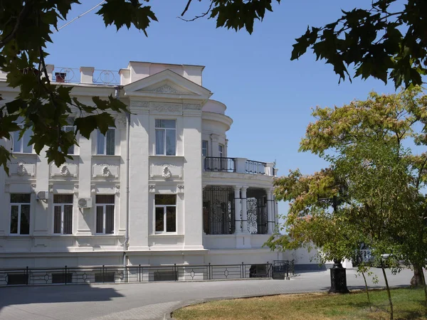 Een witte twee verdiepingen tellende landhuis, een landgoed met stucwerk molding, een balkon, bomen en een gazon tegen de achtergrond van een holle, wolkenloze hemel. Plaats van rust en toerisme... — Stockfoto