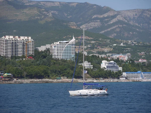 Ένα μικρό σκάφος πλέουμε το μπλε της θάλασσας κοντά στην ακτή, στρωμένο με πέτρες και τα σπίτια της πόλης που στέκεται μεταξύ του γραφικού χαρακτήρα. — Φωτογραφία Αρχείου