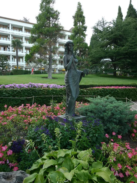 En staty av en vacker flicka i mitten av en blomma säng mitt höga gröna träd och en vit hotellbyggnaden, en fritidsgård, ett sanatorium med stora balkonger. plats för vila, Resor, turism — Stockfoto