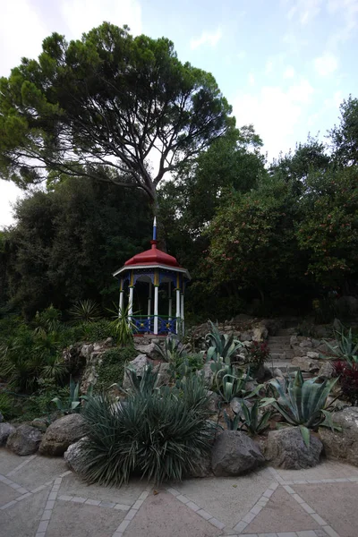 Невелика альтанка в парку з загостреним шпилем на даху, що стоїть серед великих каменів і квітів в тіні дерев . — стокове фото