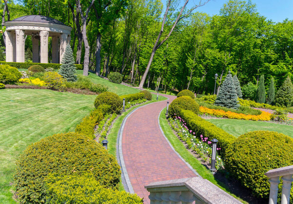 Twisted alley between beautiful flower beds. On the lawn stands a white gazebo with a black roof