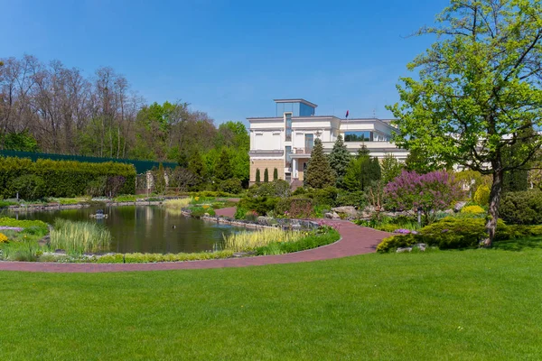 一栋两层的白色现代豪宅, 靠近绿色的草坪上, 装饰着池塘, 在蓝色晴朗的天空下保存着花坛和树木。 — 图库照片