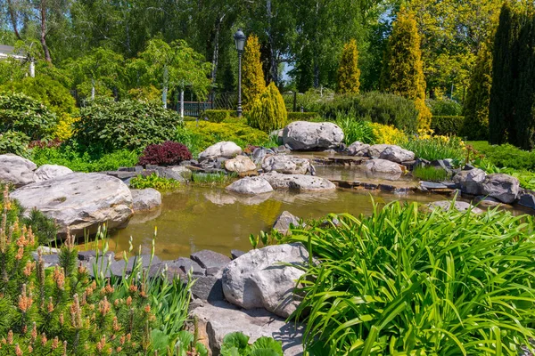 Decorative pond surrounded by boulders, flowers, green bushes, deciduous and coniferous trees — Stock Photo, Image