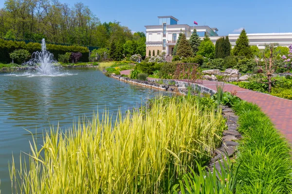 El paisaje en el parque es un día soleado brillante con la magnificencia de las plantaciones verdes de flores junto a un estanque con una fuente de árboles altos y hermosos y un edificio ligero con una bandera voladora en el techo . — Foto de Stock