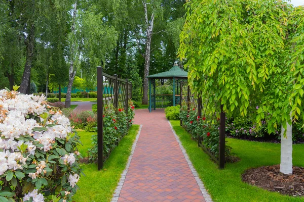 O beco do parque ao longo da cerca com rosas vermelhas que levam a um mirante decorativo com um telhado verde — Fotografia de Stock