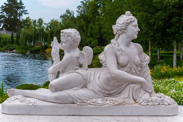 Bella scultura di una donna mezza nuda con un bambino con le ali dietro la schiena sullo sfondo di uno stagno in un parco di fiori e natura verde — Foto Stock