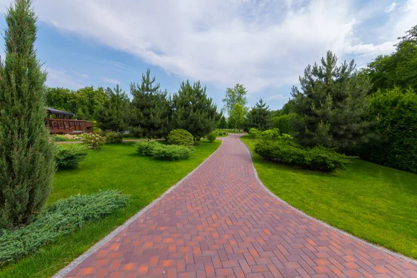 The path in the park is laid out with red tiles between green grasses and ornamental bushes. Mezhygirya Ukraine — Stock Photo, Image