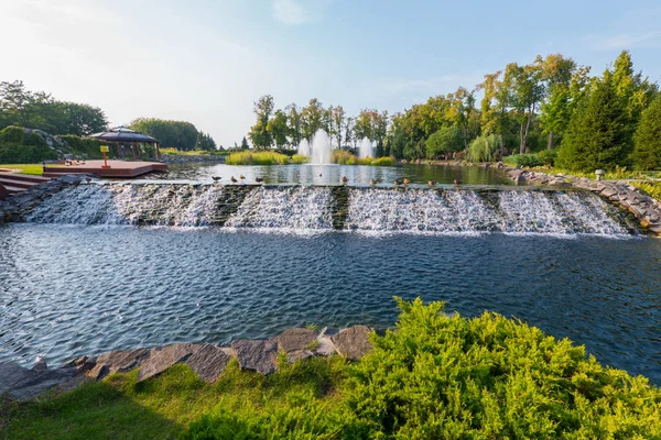 Uma cachoeira em cascata artificial flui de um lago no parque perto de um mirante de madeira com um terraço. Novi Petrivtsi, Ucrânia — Fotografia de Stock