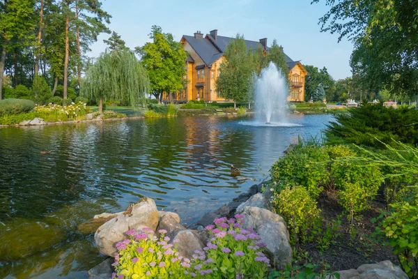 Hermoso diseño de paisaje alrededor de un lago con una fuente cerca de una casa de madera. Novi Petrivtsi, Ucrania — Foto de Stock
