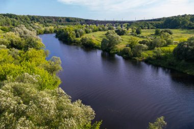Bir çayır ile uzaktan görülebilir köprü ayağı arasında akan güzel bir nehir kıyısında büyüyen yeşil ağaçlar ile pitoresk bir doğa.