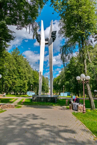 Um sinal comemorativo em um parque com pessoas descansando à sombra de uma árvore contra um céu azul com nuvens lentamente flutuantes . — Fotografia de Stock