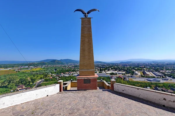 Signo conmemorativo en forma de pilar alto de piedra con un águila en la parte superior con alas extendidas. Con la ciudad debajo de ella acostada en un valle verde . — Foto de Stock