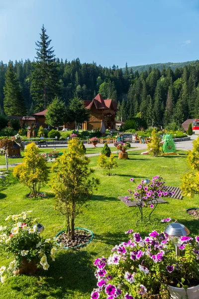 Um belo jardim com um gramado verde e flores sentadas nele com um slide de crianças e árvores jovens contra o pano de fundo de uma floresta densa e um topo de montanha que pode ser visto à distância . — Fotografia de Stock