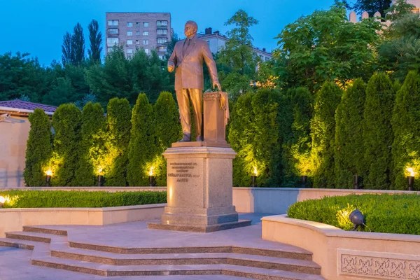 Monumento de un hombre parado en un parque sobre un pedestal. Iluminado por linternas en la oscuridad del crepúsculo de la noche con hermosos macizos de flores verdes creciendo lado a lado . — Foto de Stock