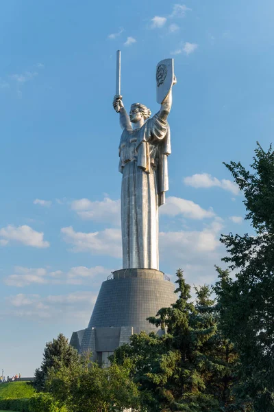 Un monumento con una alta estatua de la patria con los brazos levantados hacia arriba sosteniendo una espada en otro escudo con símbolos soviéticos de pie contra el cielo con escasas nubes . — Foto de Stock
