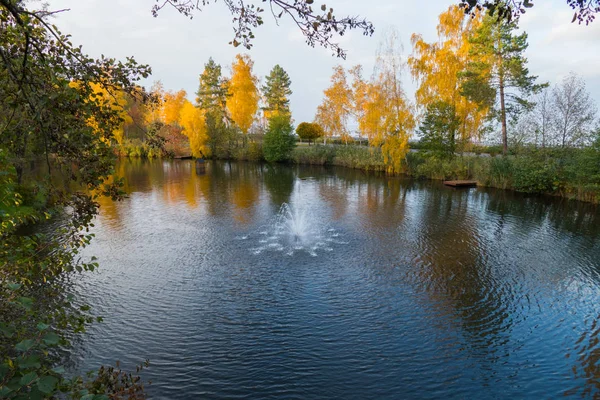 A small fountain in the middle of the pond in the early autumn with trees growing on the shore. With already yellowed on some and still green on other foliage.